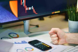 Close-up of an investor's angry fist on a desk, next to a smartphone and a computer screen showing a plummeting stock market chart, symbolizing financial collapse.