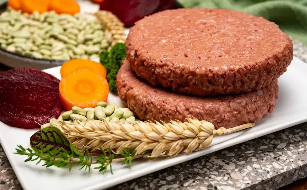 Two plant-based burger patties served on a white plate with fresh ingredients like wheat, beans, and carrots. it's synthetic meat startups.