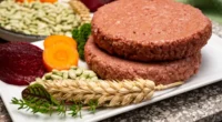 Two plant-based burger patties served on a white plate with fresh ingredients like wheat, beans, and carrots. it's synthetic meat startups.