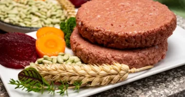 Two plant-based burger patties served on a white plate with fresh ingredients like wheat, beans, and carrots. it's synthetic meat startups.